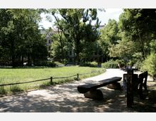 Ein kleiner, begrünter Park mit einer offenen Wiese, umgeben von Bäumen und einem geschwungenen Kiesweg. Im Vordergrund stehen Holzbänke, während im Hintergrund mehrstöckige Stadthäuser teilweise hinter dem Grün sichtbar sind.