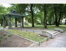 Ein kleiner Park mit einer überdachten, offenen Struktur aus Ziegelsteinpfeilern und einem grünen Dach. Umgeben von Bäumen, gibt es gepflasterte Wege, niedrige Geländer und eine breite Steintreppe im Vordergrund.