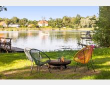 Uferwiese an einem ruhigen See mit drei farbigen Lounge-Stühlen um eine flache Feuerschale. Im Hintergrund sind Stege am Wasser sowie Häuser und Bäume am gegenüberliegenden Ufer zu sehen.