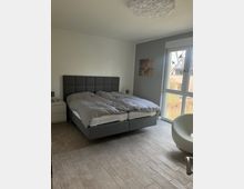 Ein modernes Schlafzimmer mit einem grauen Boxspringbett, hellen Fliesenboden und weißen Wänden. Ein großes Fenster mit Blick nach draußen lässt viel Tageslicht herein, daneben steht ein weißer Stuhl, und ein abstraktes Bild hängt an der Wand.