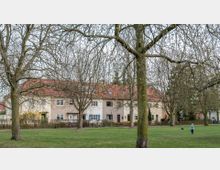 Mehrfamilienhäuser mit roten Ziegeldächern und hellen Fassaden stehen hinter einer Reihe von großen, noch kahlen Bäumen. Im Vordergrund befindet sich eine große Wiese, auf der ein Kind mit einem Ball zu sehen ist.