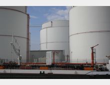 Große, weiße Tanks eines Industriegeländes, umgeben von Rohrleitungen und Anlagen. Im Vordergrund ist ein Frachtschiff mit Beladungseinrichtungen zu sehen, während der Himmel leicht bewölkt ist.