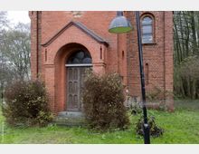 Backsteinfassade eines historischen Gebäudes mit einem hölzernen Eingangstor unter einem Rundbogen. Im Vordergrund befindet sich eine Wiese mit Büschen sowie eine schräg stehende Straßenlaterne, im Hintergrund sind Bäume sichtbar.