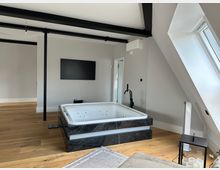 Modernes Dachgeschosszimmer mit freistehender Whirlpool-Badewanne auf dunklem Marmorsockel, Holzfußboden und sichtbaren schwarzen Deckenbalken. Rechts sorgt ein großes Dachfenster in der Schräge für Tageslicht; an der Wand hängen ein Fernseher und eine Klimaanlage.