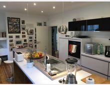 Moderne Küche mit zentraler Kochinsel, einer Spüle und Hängelampen aus Metall. An der Wand befinden sich zahlreiche gerahmte Fotos, und die Arbeitsflächen sind mit Haushaltsgeräten wie einem Mixer, einem Toaster und einer Kaffeemaschine ausgestattet.