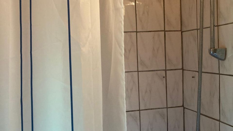 Geflieste Duschkabine mit weißen Wandfliesen und dunklen Fugen, ausgestattet mit Handbrause an einer Stange und Mischarmatur. Links hängt ein weißer Duschvorhang mit zwei blauen Längsstreifen.