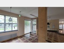 Ein Innenraum mit großen Fenstern aus Glasbausteinen und einer Wand aus gelben Ziegeln. Der Raum hat helle Wände, Hängelampen und einen gefliesten Boden in Rot- und Beigetönen, mit Blick nach draußen auf Grünflächen.