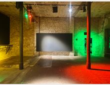 Ein leerer Raum mit grob verputzten Backsteinwänden, einer dunklen Tafel in der Mitte und Betonboden, beleuchtet von Scheinwerfern in grünen, roten und gelben Farben. Zwei Metallpfeiler teilen den Raum; rechts ist eine beleuchtete Tür erkennbar.