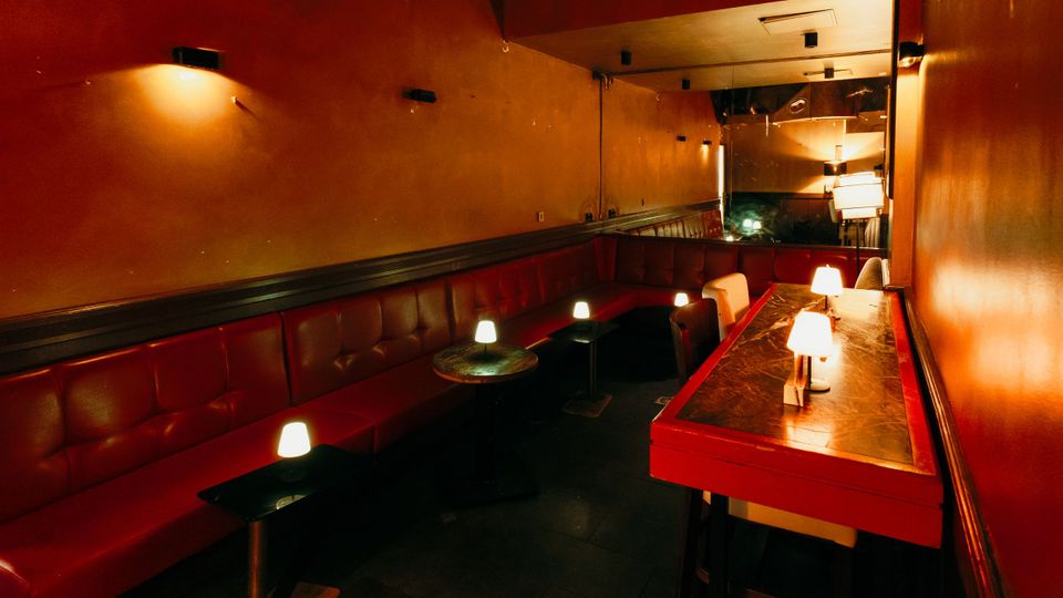 Schmaler Lounge- oder Barbereich mit langen roten Leder-Sitzbänken entlang der Wand, kleinen runden und eckigen Tischen sowie gedämpften Tischlampen. Warme, orangefarbene Wände und ein langer Tresen mit marmorierter Oberfläche prägen den Innenraum.