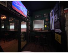 Gemütlicher Bar- oder Pub-Innenraum mit dunklen Holzvertäfelungen, hohen Barhockern und einem Wandfernseher über einem Spiegelbereich. An den Wänden hängen Bilder und Bierschilder; ein Fenster mit Jalousie sowie ein geöffnetes Fenster mit Läden prägen die Rückwand.