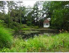 Ein grüner Garten mit einem kleinen Teich, umgeben von Büschen und Bäumen. Im Hintergrund steht ein weißer Pavillon mit roten Dachziegeln und runden Fenstern.