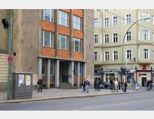 Straßenszene mit einem mehrstöckigen Gebäude aus Beton und Ziegelplatten links sowie einem sanierten Wohnhaus mit heller Fassade rechts. Im Erdgeschoss des rechten Gebäudes befindet sich eine Apotheke, während Passanten und Radfahrer den Gehweg entlanggehen und an einer Straßenkreuzung warten.