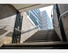 Treppenaufgang aus einem U-Bahnhof mit seitlichen Metallgeländern und Steinwänden, im Hintergrund sind moderne Bürogebäude mit rechteckigen Fenstern sichtbar. Helles Tageslicht fällt durch die Öffnung am oberen Ende der Treppe.