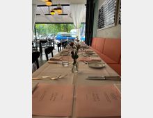 Ein modern eingerichtetes Restaurant mit gedecktem Tisch entlang einer orangen Sitzbank. Auf dem Tisch stehen Wassergläser, Besteck, rosa Menükarten und eine kleine Vase mit Blumen; im Hintergrund sind große Fenster mit weißen Vorhängen und Blick auf geparkte Autos und Bäume.