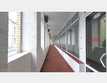 Ein langer, überdachter Gang mit weißen Backsteinwänden und großen Glasfenstern auf der rechten Seite, auf denen "Eingang" in roter Schrift steht; der Boden ist mit roten Fliesen belegt. Links sind verputzte Mauerabschnitte mit Blick auf die Straße durch schmale Fenster erkennbar.