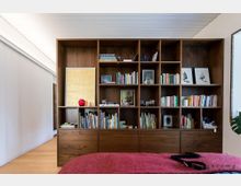Holzregalwand in einem modernen Wohnraum mit mehreren Fächern, gefüllt mit Büchern, gerahmten Bildern, Dekorationsgegenständen und persönlichen Objekten; davor ein Bett mit rotem Überwurf. Der Boden besteht aus Holz, und die Decke ist weiß mit Lamellenstruktur.