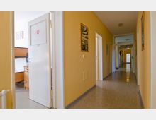 Ein langer, schmaler Flur mit gelb gestrichenen Wänden und dunklem Bodenbelag; an der linken Seite befindet sich eine offene Tür, die in ein Zimmer mit Holzmöbeln und einem Fernseher führt. Bilder hängen rechts und links an den Wänden, und am Ende des Flurs ist eine weitere Tür sichtbar.