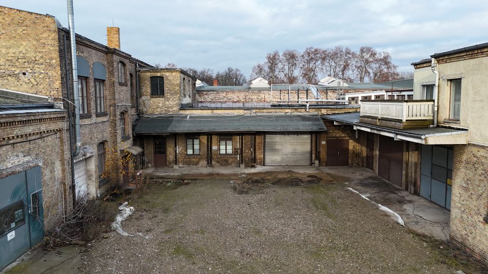 Innenhof eines alten Industrie- oder Lagerkomplexes mit Backsteinfassaden, niedrigen Anbauten und mehreren Metalltoren. Der unbefestigte Hof ist überwiegend kiesbedeckt, umgeben von Flachdächern, Rohren und einer kleinen Balkonterrasse; im Hintergrund stehen kahle Bäume.