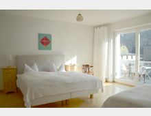 Helles Schlafzimmer mit weißer Einrichtung, einem Doppelbett mit gepolstertem Kopfteil, zwei holzfarbenen Nachttischen und einer Glas-Balkontür, die auf eine Terrasse mit Tisch und Stühlen führt. An der Wand hängt ein abstraktes Kunstwerk in Rot und Grün.