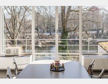 Großer Besprechungsraum mit einem Tisch und modernen Stühlen vor einer raumhohen Fensterfront, die den Blick auf einen Fluss, Bäume und eine Brücke in einer parkartigen Umgebung freigibt.