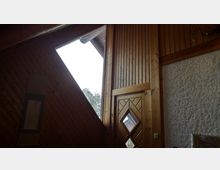 Holzvertäfelter Innenraum mit einer schrägen Wand und einem dreieckigen Fenster, das Tageslicht hereinlässt. Rechts unten ist eine Holztür mit einem rautenförmigen Glaseinsatz, und daneben eine Wand mit grober Putzstruktur.