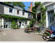 Ein Innenhof mit gepflastertem Boden und zwei Gebäuden im Hintergrund. Das linke Haus hat eine Fassade mit Fenstern, Türen und Kletterpflanzen, während am rechten Gebäude eine Treppe mit Blumenbeeten und Topfblumen aufgestellt ist. Ein Holzpicknicktisch steht zentral im Hof.