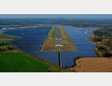 Luftaufnahme eines großen Solarparks mit einer stillgelegten Landebahn in der Mitte. Die Fläche ist von dicht angeordneten Reihen von Solarmodulen umgeben und befindet sich in einer ländlichen, bewaldeten Umgebung.