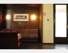 Innenraum einer Gaststätte oder eines Cafés mit dunkler Holzvertäfelung und braunen Polstermöbeln. An der Wand hängen Wandlampen und ein gerahmtes Bild; rechts befindet sich eine Glastür, daneben ein Nichtraucherschild.