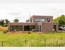 Modernes Einfamilienhaus mit flachem Dach, rechteckigen Formen und einer Fassade in erdigen Brauntönen. Das Gebäude verfügt über große Fensterfronten, eine Terrasse und einen gepflegten Garten im Vordergrund.