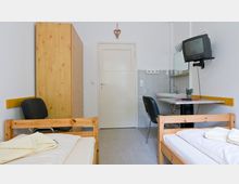 Ein einfacher Raum mit zwei Einzelbetten aus Holz, einem Kleiderschrank, einem kleinen Waschbecken mit Wandspiegel, einem klappbaren Tisch mit Stuhl und einem alten Röhrenfernseher, der an der Wand montiert ist. Die Wände sind weiß, und der Boden besteht aus grauem Linoleum.