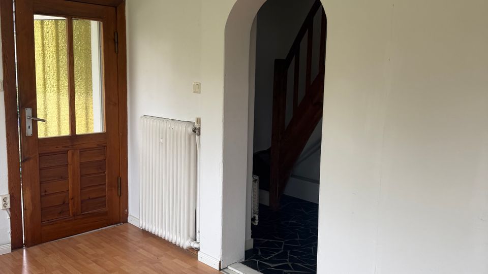 Leerer Eingangsraum mit hellem Laminatboden und weißen Wänden, links eine Holz-Eingangstür mit Glasfüllung. Daneben ein Heizkörper und ein Rundbogen-Durchgang in einen Flur mit dunklem Boden, von dem eine Holztreppe nach oben führt.