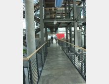 Ein moderner Innenbereich mit einem erhöhten Gehweg, der von Metallgeländern mit Holzhandläufen gesäumt ist. Im Hintergrund sind Stahlträger, Treppen und Glaswände sichtbar, die eine offene und industrielle Architektur zeigen.