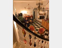 Innenansicht eines gemütlichen Wohnzimmers mit kariertem Fußboden, einem Kronleuchter und dekorierten Wänden. Im Mittelpunkt sitzen zwei Personen, eine spielt Gitarre, die andere eine Trommel, während mehrere Sessel um einen kleinen Tisch gruppiert sind.