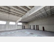 Ein leerer, industrieller Innenraum mit Sichtbetonwänden, einem flachen Balkendeckenaufbau und großen Fenstern, die diffuses Licht hereinlassen. Der Boden weist eingelassene Schienen und versetzte Glasplatten auf, die Umgebung wirkt unbehandelt und roh.