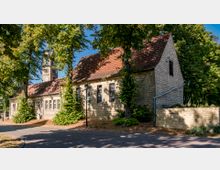 Steingebäude mit rotem Satteldach, flankiert von Bäumen, im Vordergrund ein gepflasterter Weg; im Hintergrund ein Kirchturm mit Uhr und gotischen Fenstern.