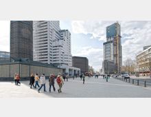 Städtischer Platz mit Fußgängern, umgeben von modernen Hochhäusern und einem niedrigen Gebäude mit Glasbausteinfassade; im Hintergrund ein im Bau befindlicher Turm und bewölkter Himmel.