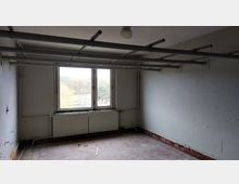 Leerer Raum mit weißen Wänden, sichtbaren Heizungsrohren und einem heruntergekommenen Bodenbelag aus Fliesen. Die Decke weist mehrere Metallstangen auf, und durch ein Doppelfenster ist eine herbstliche Landschaft zu sehen.