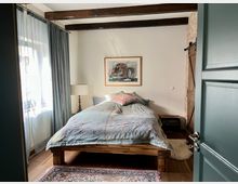 Schlafzimmer mit Holzboden und sichtbaren Holzdeckenbalken, ausgestattet mit einem Holzbett, farbiger Bettwäsche und einem Nachttisch. Im Raum befinden sich ein großer Vorhang, eine Stehlampe und eine Schiebetür aus Holz vor einer Wand in Ziegeloptik.