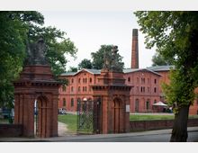 Backsteinbau im Industriestil mit dekorativem Eingangstor aus rotem Backstein und Skulpturen verziert. Im Hintergrund sind weitere Backsteingebäude, ein Schornstein und Grünflächen zu sehen.