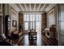 Elegante Halle im historischen Stil mit Kassettendecke, gemusterten Wänden und schwarz-braunem Fliesenboden. In der Mitte steht ein Holztisch mit Getränkeflaschen vor hohen verglasten Türen; seitlich sind ein Ledersofa, eine Standuhr sowie ein Kamin mit Blumendekoration zu sehen.