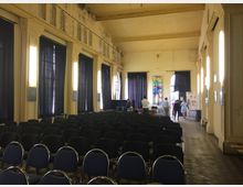 Innenansicht eines alten Versammlungssaals mit hohen Decken, gelb-beigen Wänden und hohen Fenstern, die mit schwarzen Vorhängen bedeckt sind. Reihen schwarzer und blauer Stühle sind aufgestellt, und im Hintergrund sind ein bunter Wandteppich sowie einige Personen in der Nähe einer geöffneten Tür zu sehen.