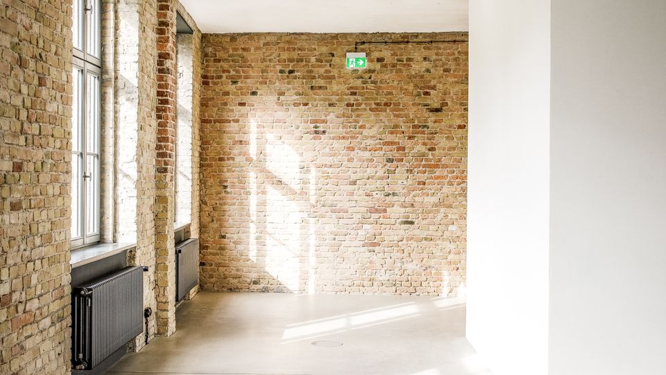 Heller, leerer Innenraum im Loft-Stil mit freiliegenden Backsteinwänden, glatter Betonboden und großer Fensterfront links. Sonnenlicht fällt in den Raum; an der Rückwand ist ein grünes Notausgangsschild sichtbar.
