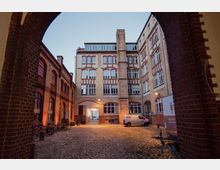 Ein gepflasterter Innenhof, umgeben von historischen Backsteingebäuden mit großen Fenstern und roten Zierelementen. Im Hof stehen geparkte Fahrräder und Lieferwagen, und der Eingang ist von einem Torbogen aus Backstein eingerahmt.