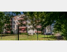 Mehrstöckiges Wohnhaus mit Balkonen in einer grünen Umgebung. Im Vordergrund befinden sich eine Wiese, Bäume und geparkte Autos, die einen ruhigen Wohnbereich andeuten.