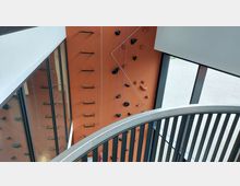Ein Indoor-Kletterbereich mit einer orangenen Kletterwand, ausgestattet mit Klettergriffen, Trittstangen und Seilen. Im Vordergrund ist ein geschwungenes Treppengeländer, im Hintergrund große Fenster mit Blick nach draußen.