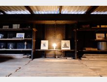 Holzregale in einem rustikalen Innenraum mit Metallwänden, darauf Bücher, Bilderrahmen und Dekorationsgegenstände. Eine kleine Tischlampe beleuchtet zwei gerahmte Drucke mit Motiven von Segelschiffen in der Mitte.