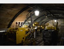 Ein unterirdischer Bergwerksstollen mit gewölbter Decke aus Holz und Metallstreben, beleuchtet durch Käfiglampen. Im Vordergrund sind gelbe Bergbaumaschinen und ein schwarzer Förderwagen auf Gleisen zu sehen, die von Steinwänden umgeben sind.