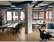 Ein modernes Großraumbüro mit hellem Holzboden, mehreren weißen Schreibtischen, schwarzen Bürostühlen und Schreibtischlampen. Große Fenster mit roten Rahmen lassen viel Tageslicht herein, während die Decke mit runden Schallschutzplatten ausgestattet ist.