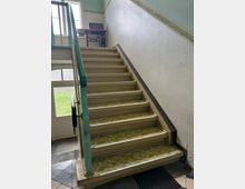 Innenbereich eines Treppenhauses mit einer beige lackierten Holztreppe und gemusterten Stufen. Rechts und links sind einfache Handläufe, im Hintergrund ein Fenster sowie ein kleiner Holztisch mit einer alten Näheinrichtung.