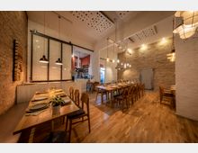 Gemütlich eingerichtetes Restaurant mit Holztischen und -stühlen, freigelegten Ziegelwänden und moderner Hängelampenbeleuchtung. Im Hintergrund ist eine offene Küche sowie eine Tür mit industriellem Design zu sehen.